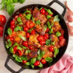 Overhead view of a pan with tomato basil chicken thighs surrounded by a linen napkin, shallots, basil, and tomatoes.