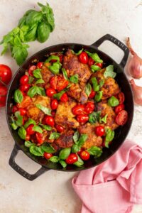 Overhead view of a pan with tomato basil chicken thighs surrounded by a linen napkin, shallots, basil, and tomatoes.