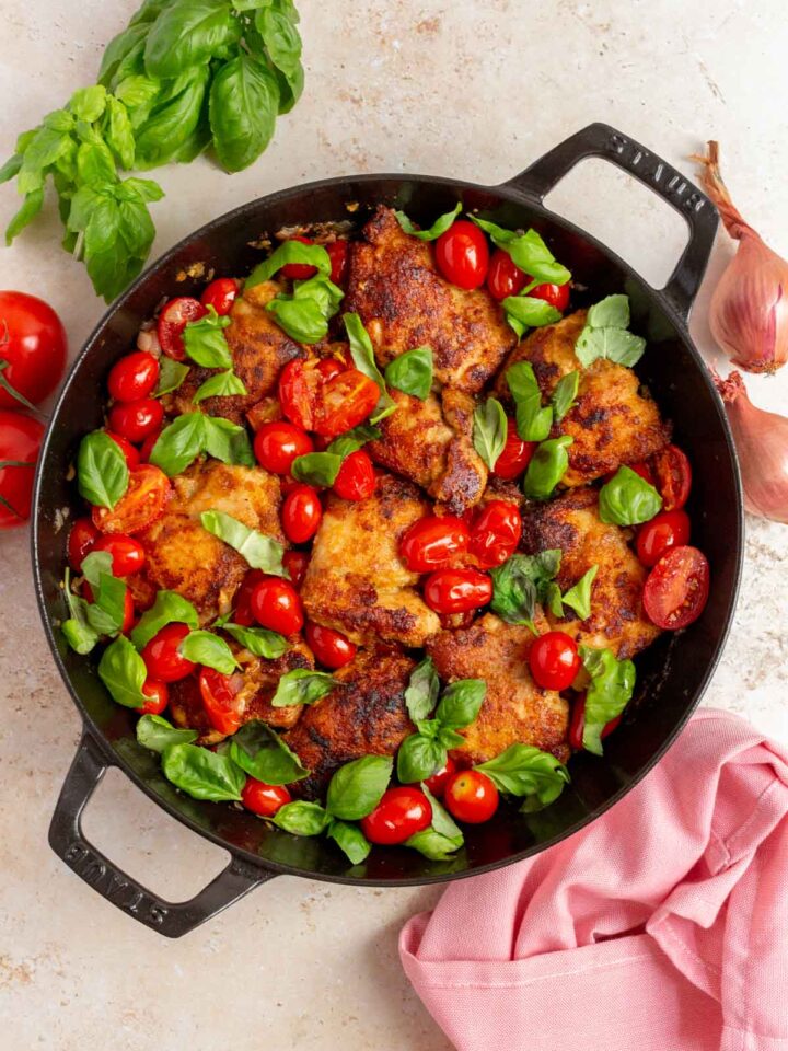 Overhead view of a pan with tomato basil chicken thighs surrounded by a linen napkin, shallots, basil, and tomatoes.