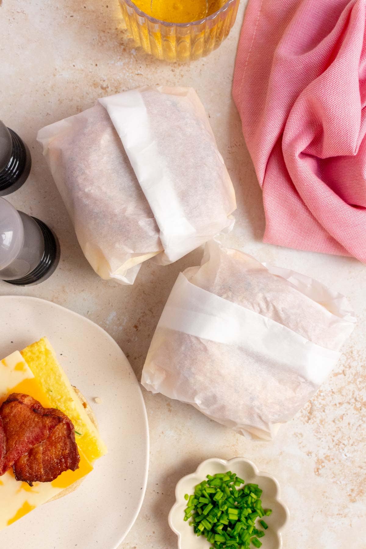 Two parchment wrapped bacon, egg, and cheese bagel with ingredients on the side.