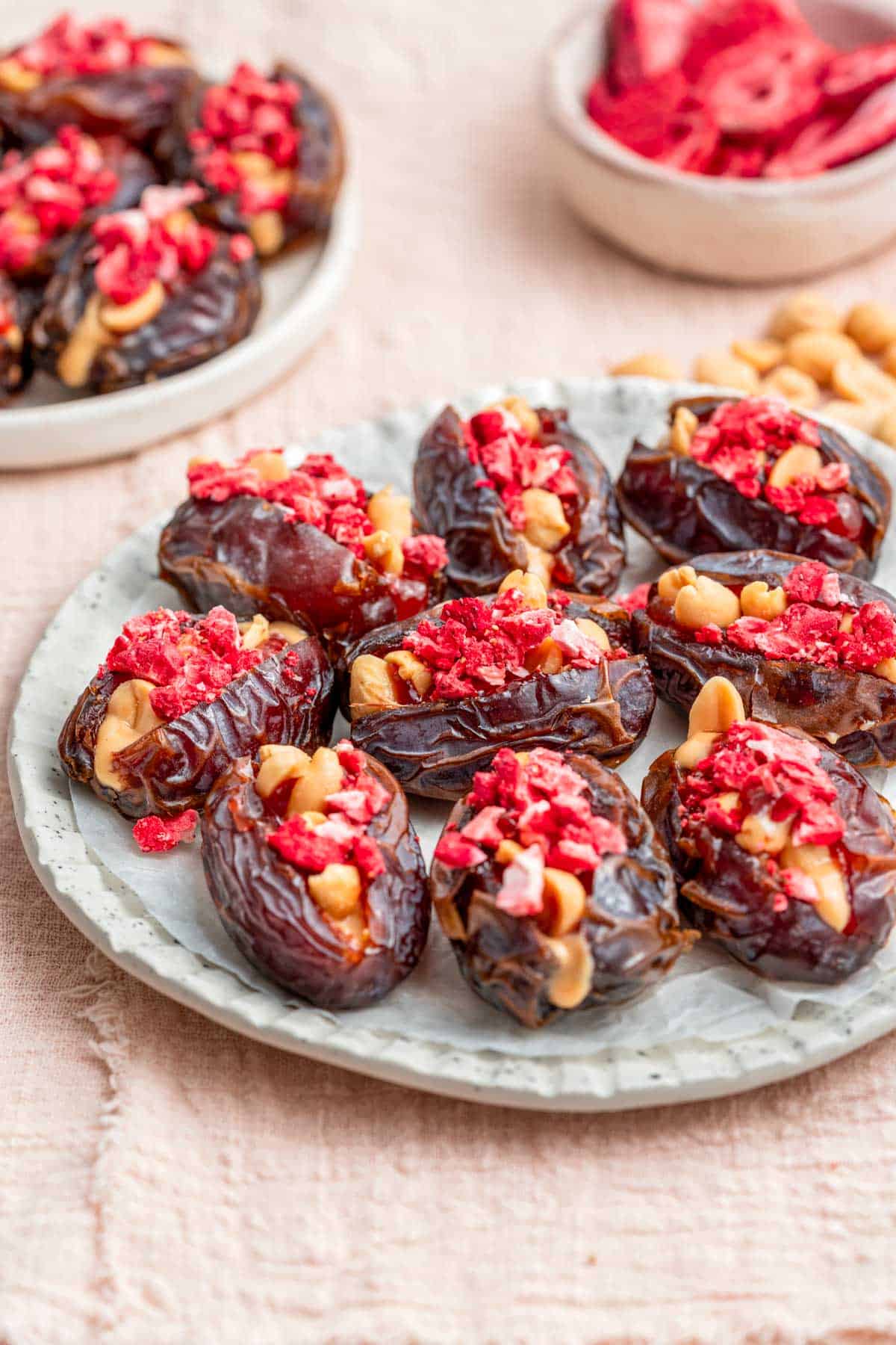An angled view of a plate of peanut butter and jelly stuffed dates. More in the background along with a bowl of freeze-dried strawberries and peanuts.