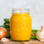 A mason jar of veggie packed broth on a wooden board surrounded by veggies.