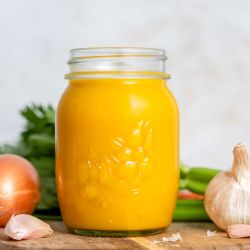 A mason jar of veggie packed broth on a wooden board surrounded by veggies.