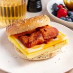 A slightly side view of a bacon, egg, and cheese bagel on a plate. Apple juice in the back along with a plate of fruit.