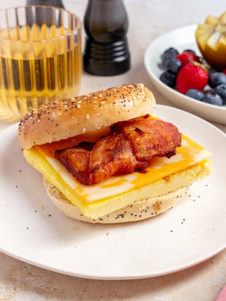 A slightly side view of a bacon, egg, and cheese bagel on a plate. Apple juice in the back along with a plate of fruit.
