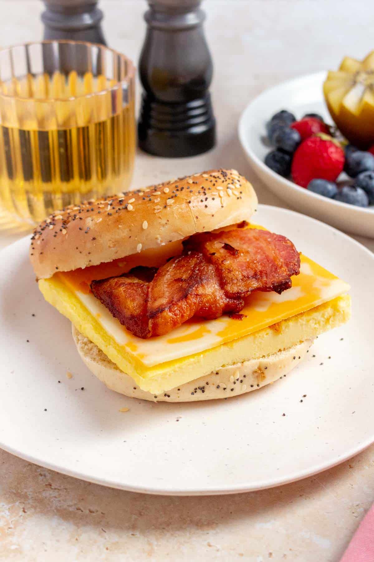 A slightly side view of a bacon, egg, and cheese bagel on a plate. Apple juice in the back along with a plate of fruit.