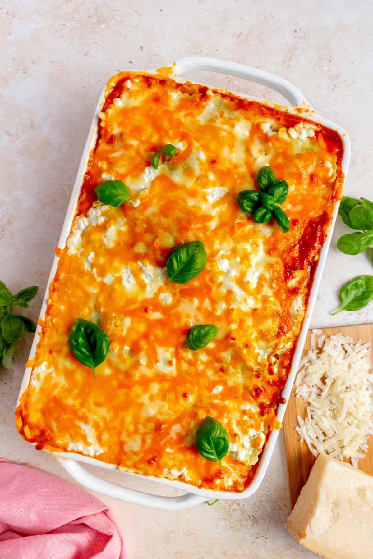 A baking dish of ravioli lasagna topped with fresh basil leaves. Parmesan on the side.