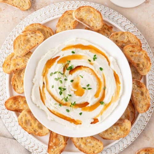 Overhead view of a bowl of whipped goat cheese with drizzles of hot honey and fresh thyme leaves on top of a plate with crostini.