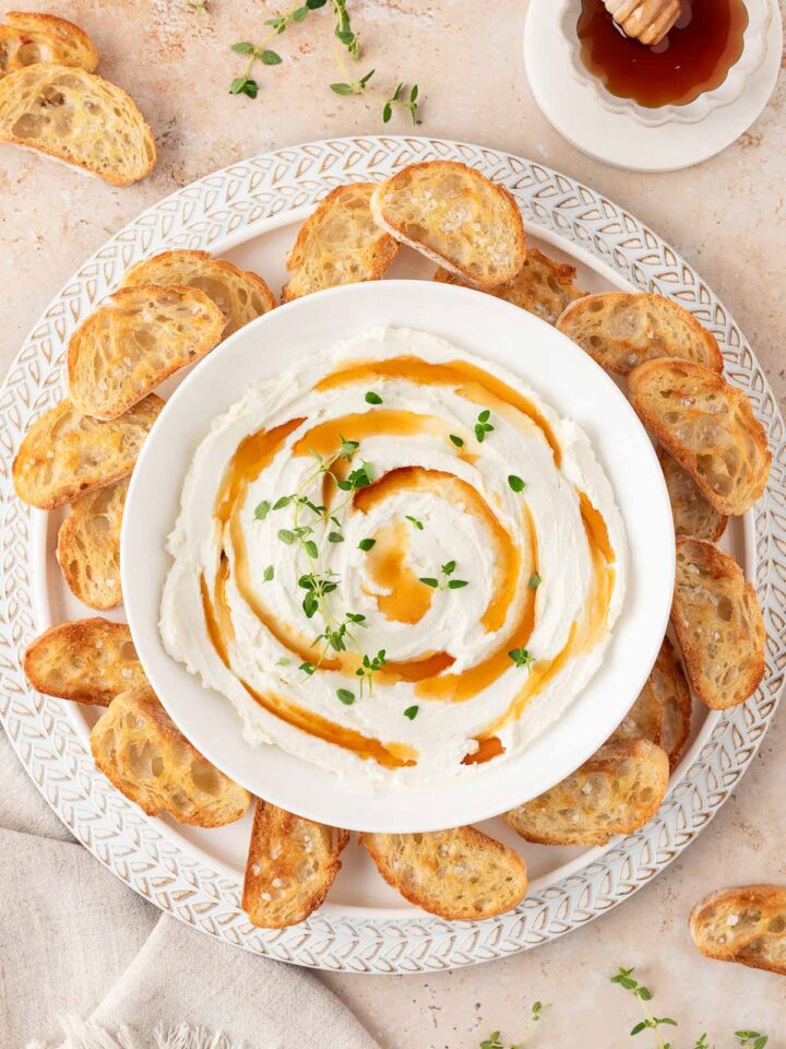 Overhead view of a bowl of whipped goat cheese with drizzles of hot honey and fresh thyme leaves on top of a plate with crostini.