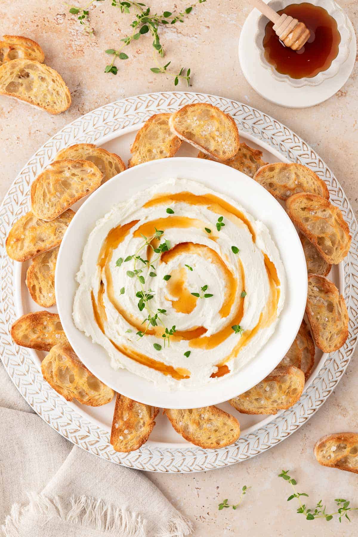 Overhead view of a bowl of whipped goat cheese with drizzles of hot honey and fresh thyme leaves on top of a plate with crostini.