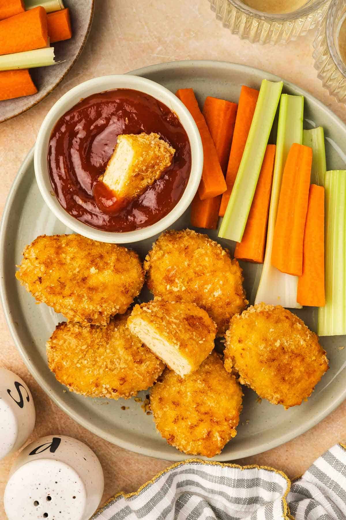 A plate with cheesy chicken nuggets, celery, and carrots. One nugget cut in half with half in a bowl of ketchup.
