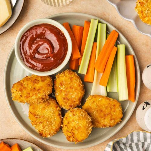 Overhead view of a plate with cheesy chicken nuggets, celery, carrots, and a bowl of ketchup.
