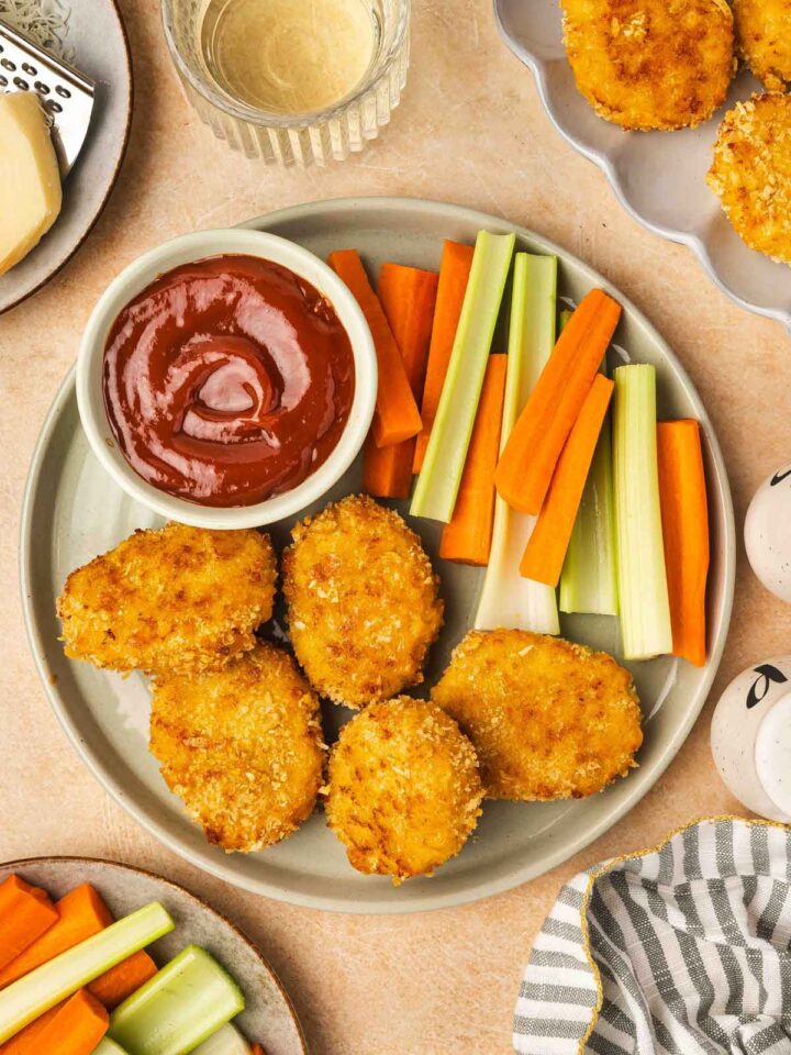 Overhead view of a plate with cheesy chicken nuggets, celery, carrots, and a bowl of ketchup.