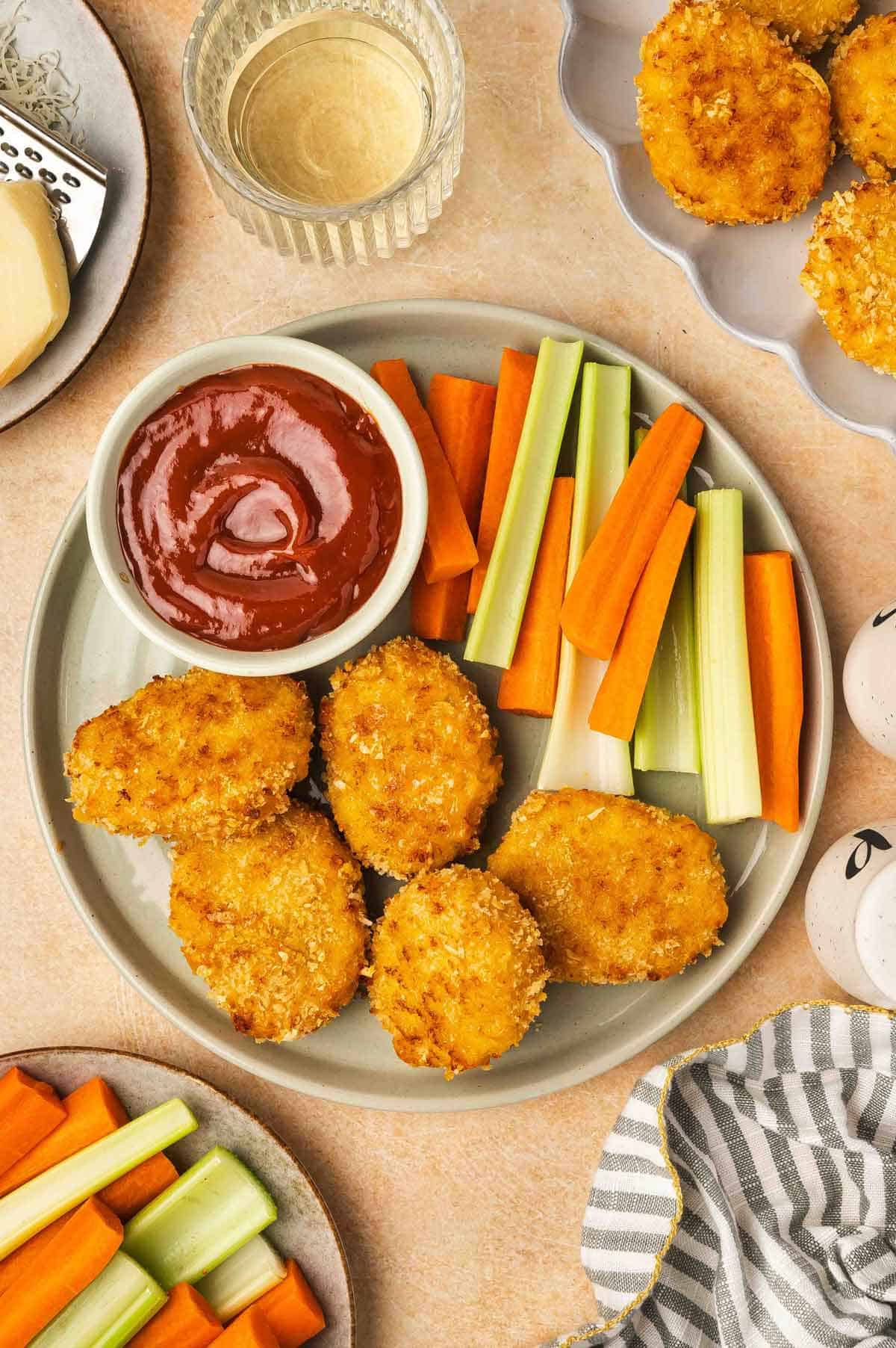 Overhead view of a plate with cheesy chicken nuggets, celery, carrots, and a bowl of ketchup.