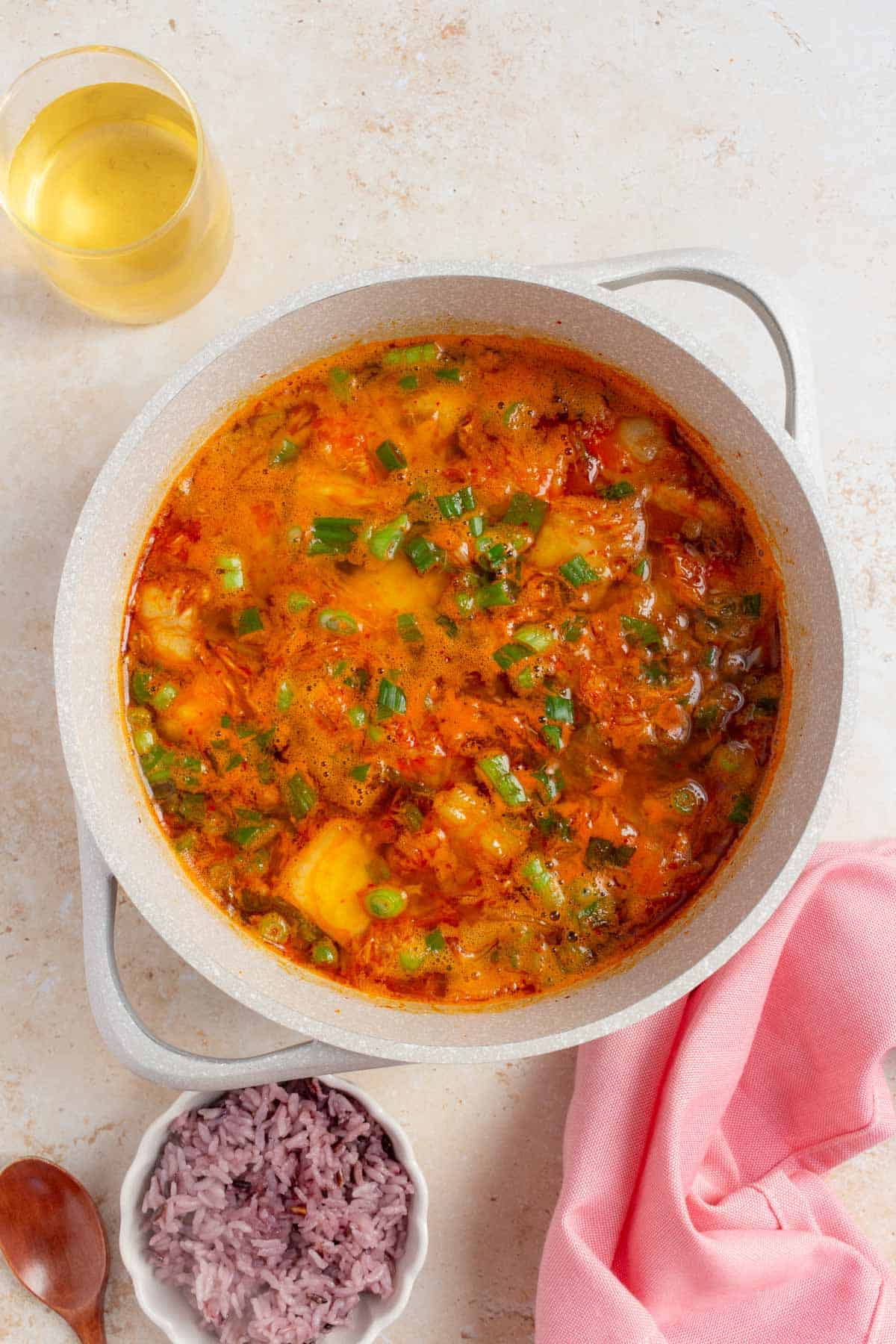 An overhead view of a large pot of tuna kimchi stew topped with green onions. A bowl of rice and drink on the side.