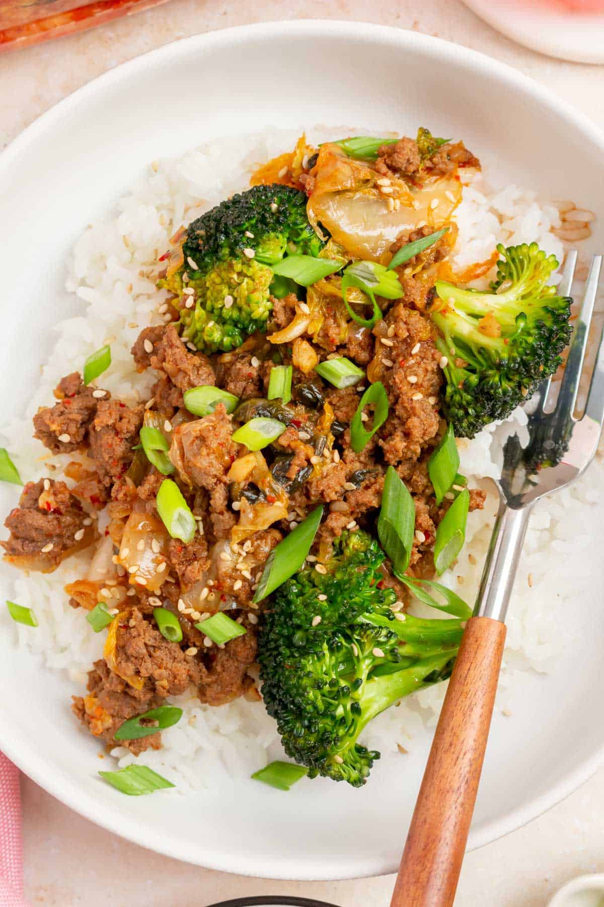 Close up view of gochujang beef and broccoli and kimchi over rice with sesame seeds and green onions with a fork.