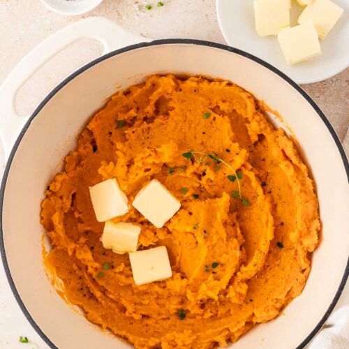 A white dutch oven with mashed sweet potatoes with knobs of butter and fresh thyme as garnish.