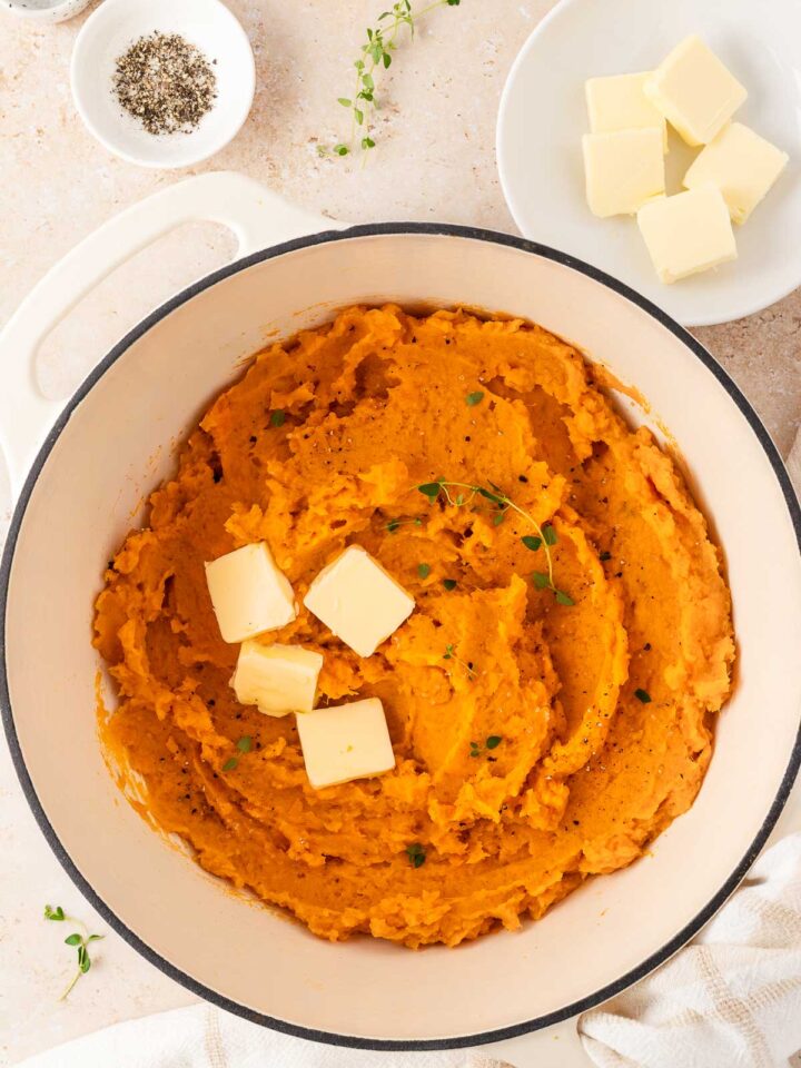 A white dutch oven with mashed sweet potatoes with knobs of butter and fresh thyme as garnish.