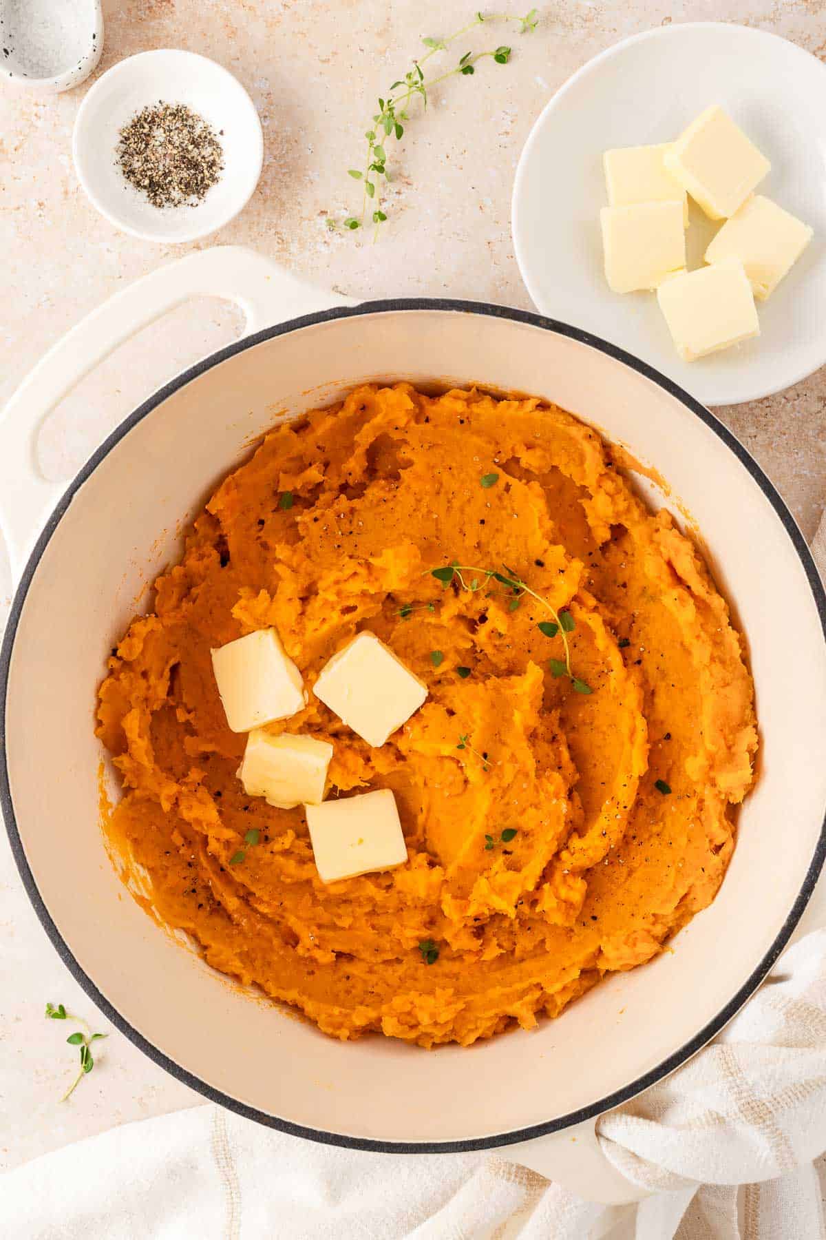 A white dutch oven with mashed sweet potatoes with knobs of butter and fresh thyme as garnish.