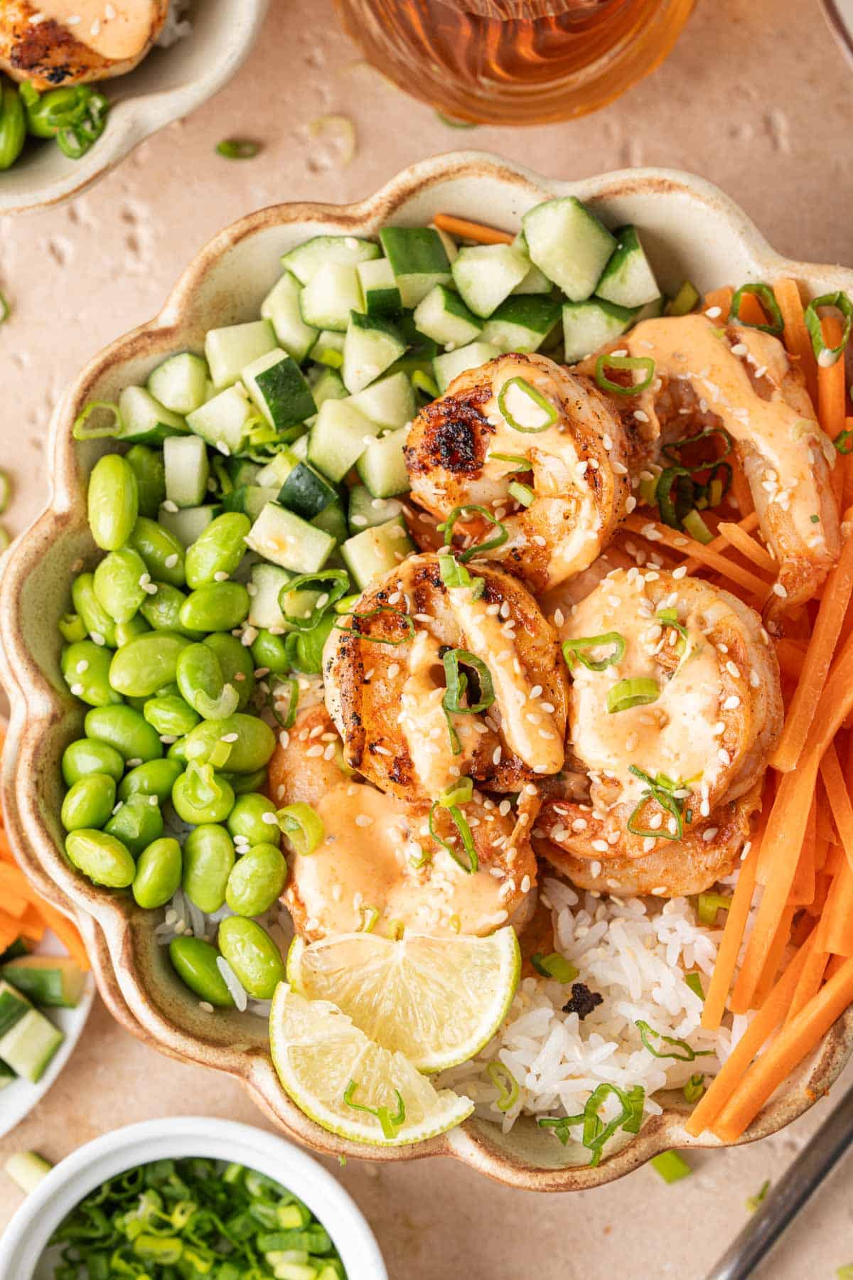 Overhead view of a bowl of shrimp rice bowls with spicy mayo with edamame, carrots, cucumbers, lime, green onions, and sesame seeds.