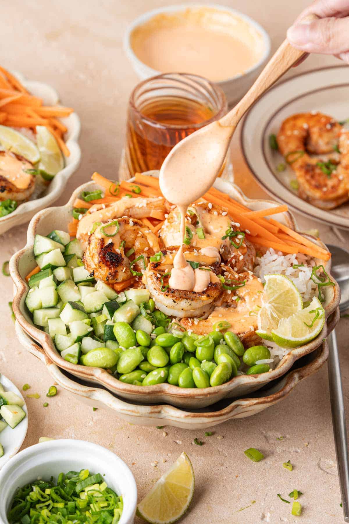 A spoon drizzling homemade spicy mayo onto a shrimp rice bowls with rice, cucumber, edamame, and carrots.