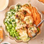 Overhead view of a bowl of shrimp rice bowls with spicy mayo with edamame, carrots, cucumbers, lime, green onions, and sesame seeds.