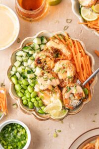 Overhead view of a bowl of shrimp rice bowls with spicy mayo with edamame, carrots, cucumbers, lime, green onions, and sesame seeds.