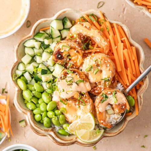 Overhead view of a bowl of shrimp rice bowls with spicy mayo with edamame, carrots, cucumbers, lime, green onions, and sesame seeds.
