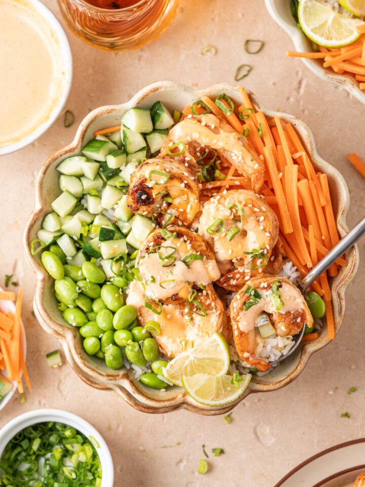 Overhead view of a bowl of shrimp rice bowls with spicy mayo with edamame, carrots, cucumbers, lime, green onions, and sesame seeds.